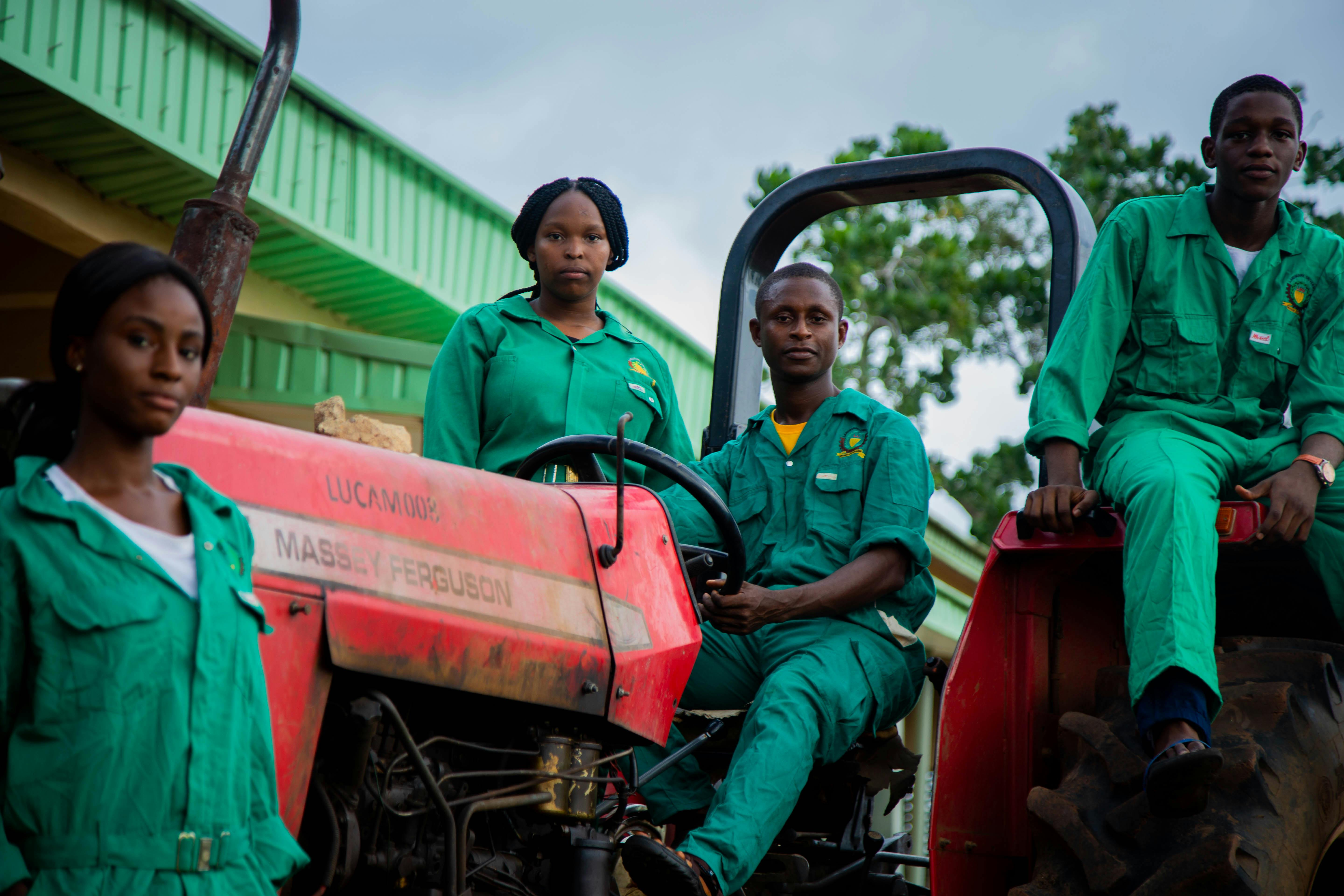 Group of young agricultural workers in green uniforms sitting on a red tractor outdoors.